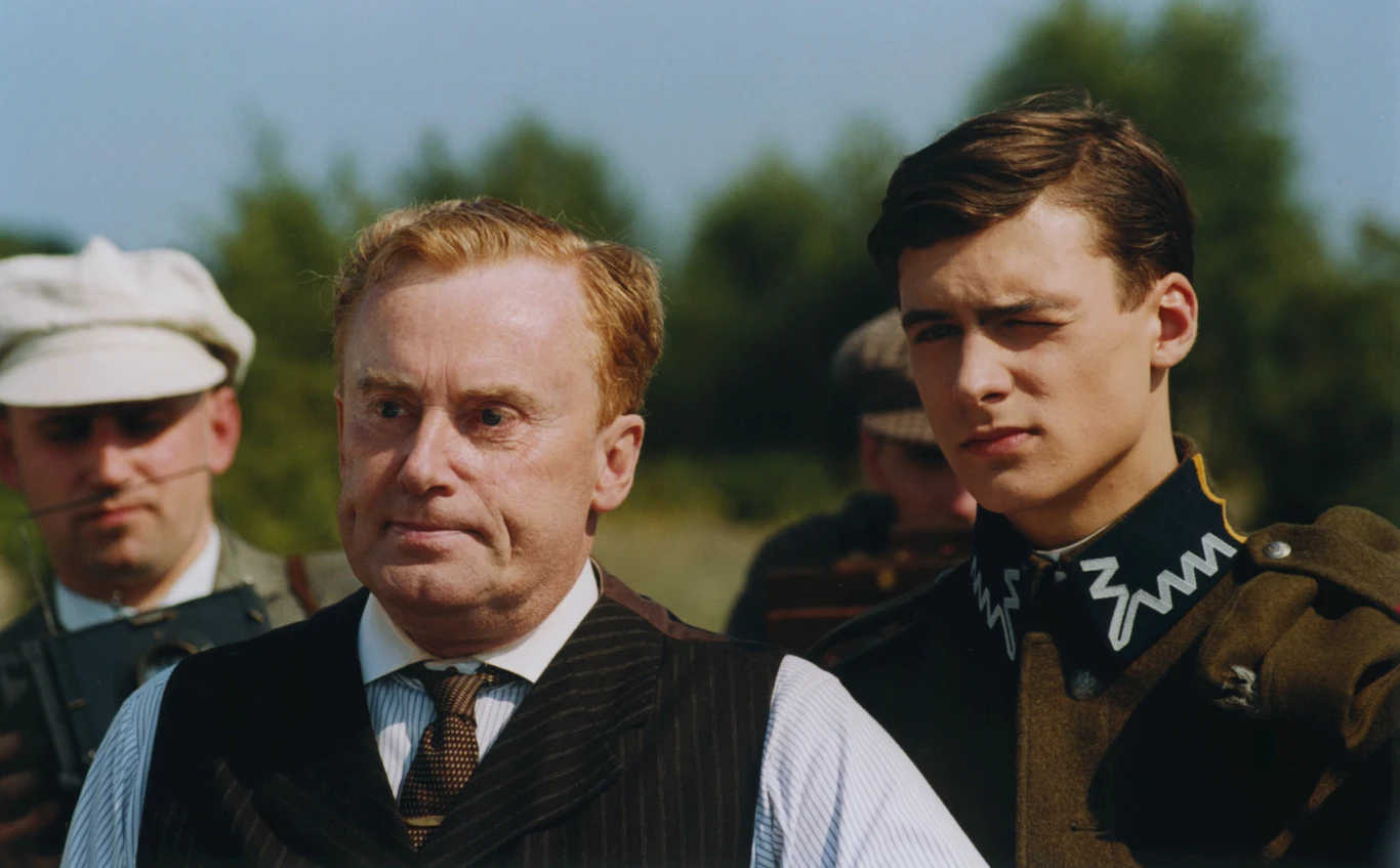 Daniel Olbrychski i Mateusz Damięcki w filmie "Przedwiośnie" Mężczyzna w średnim wieku w garniturze stoi obok młodego mężczyzny w mundurze wojskowym na tle zieleni, w tle widoczne są jeszcze inne osoby.
