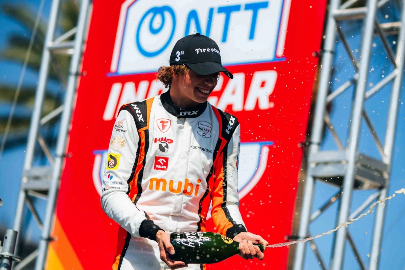 W swoim debiucie w Indy NXT Tymek Kucharczyk wywalczył miejsce na podium Sportowiec w kombinezonie wyścigowym i czapce z daszkiem świętuje zwycięstwo, trzymając butelkę szampana, z której tryska płyn. W tle widoczne elementy scenografii i logo sponsora.