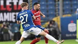 Dwóch piłkarzy walczy o piłkę podczas meczu piłkarskiego na stadionie, jeden ubrany w niebieską koszulkę z numerem 27, drugi w czerwoną koszulkę, intensywna rywalizacja na tle trybun.