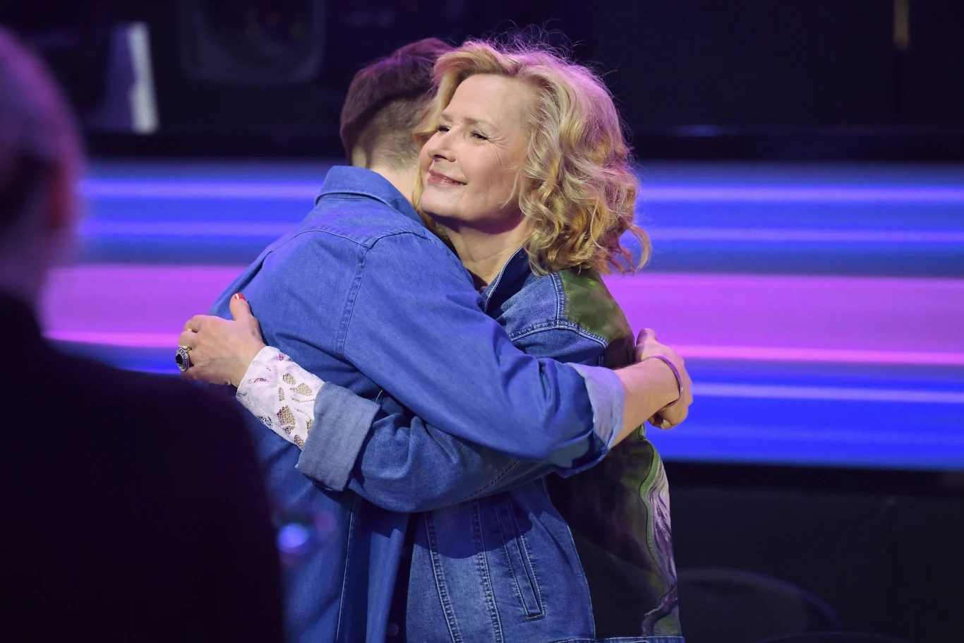 Dwie osoby w dżinsowych kurtkach obejmują się serdecznie na tle jasnych, kolorowych świateł scenicznych. Kobieta uśmiecha się z zamkniętymi oczami, wyrażając radość i wzruszenie.