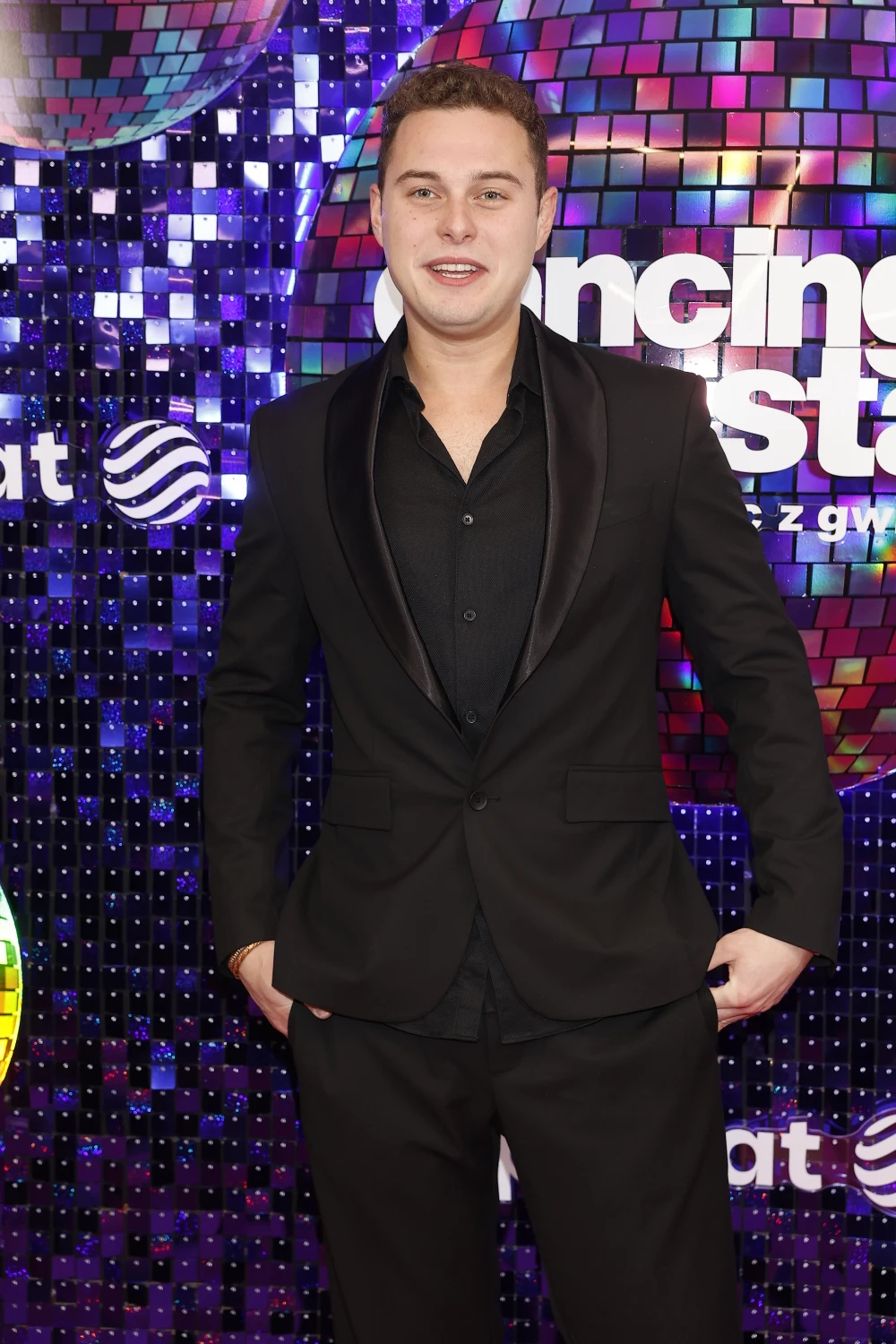 Młody mężczyzna w eleganckim, czarnym garniturze stoi na tle kolorowej ściany z cekinów oraz logotypu programu rozrywkowego z motywem tanecznym.