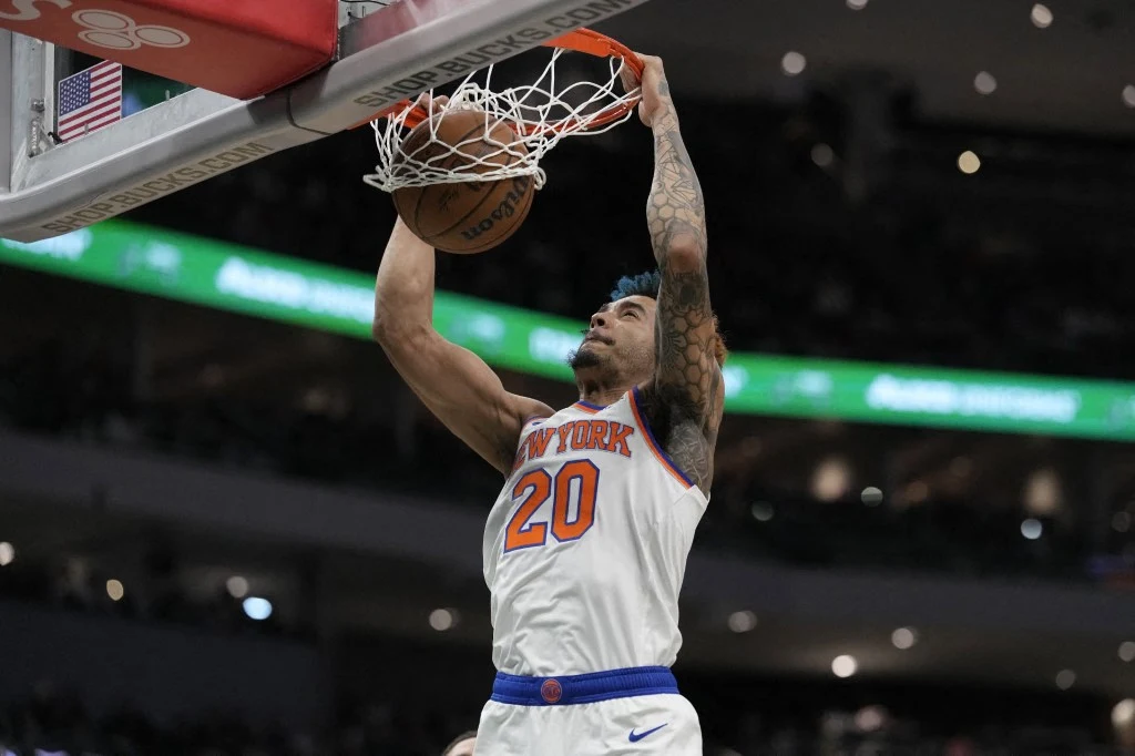 Koszykarz drużyny New York Knicks wykonuje wsad do kosza podczas rozgrywki na hali sportowej. Na pierwszym planie widać wysiłek zawodnika oraz szczegóły koszulki z numerem 20.