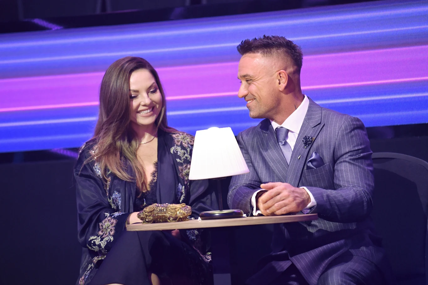 Dwie elegancko ubrane osoby, mężczyzna i kobieta, siedzą razem przy stoliku z niewielką lampką. Oboje uśmiechają się do siebie, tworząc przyjazną, intymną atmosferę. W tle widoczna jest kolorowa, świetlna scenografia.