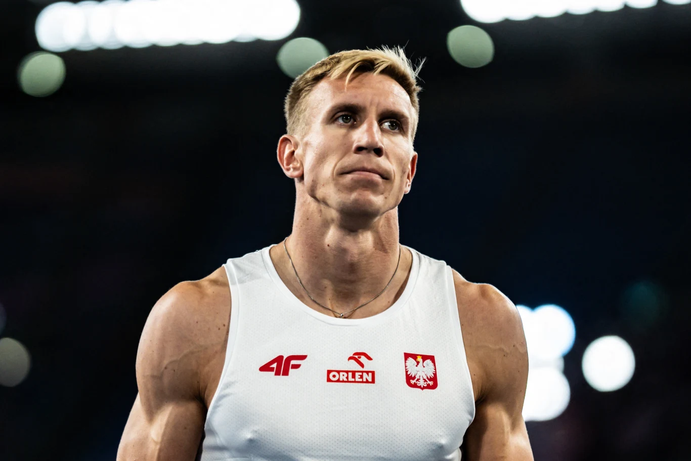 Piotr Lisek Muskularny sportowiec w białej koszulce z polskimi emblematami oraz logotypami sponsorów, na tle rozmytego stadionowego oświetlenia, z poważnym wyrazem twarzy.