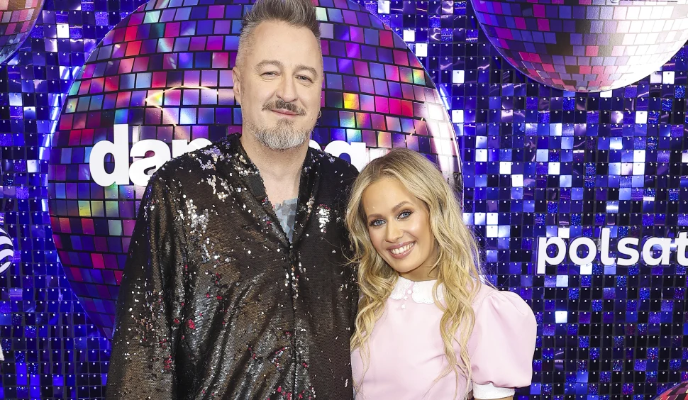 Piotr Kędzierski i Magda Tarnowska Uśmiechnięty mężczyzna w błyszczącym czarnym stroju i kobieta w różowo-białej sukience stoją razem na tle stylizowanym na ścianę z lustrzanych dyskotekowych kul i logo programu telewizyjnego.