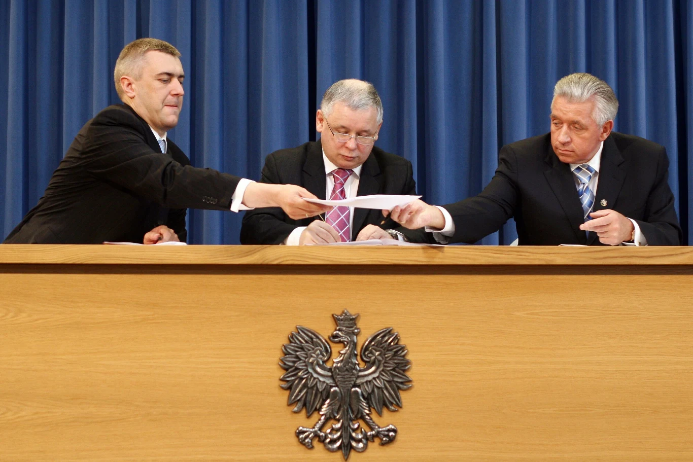 Trzech mężczyzn w garniturach siedzi za drewnianym stołem, na którego froncie widnieje godło Polski. Mężczyzna po lewej przekazuje dokument mężczyźnie po prawej. W tle niebieska zasłona.