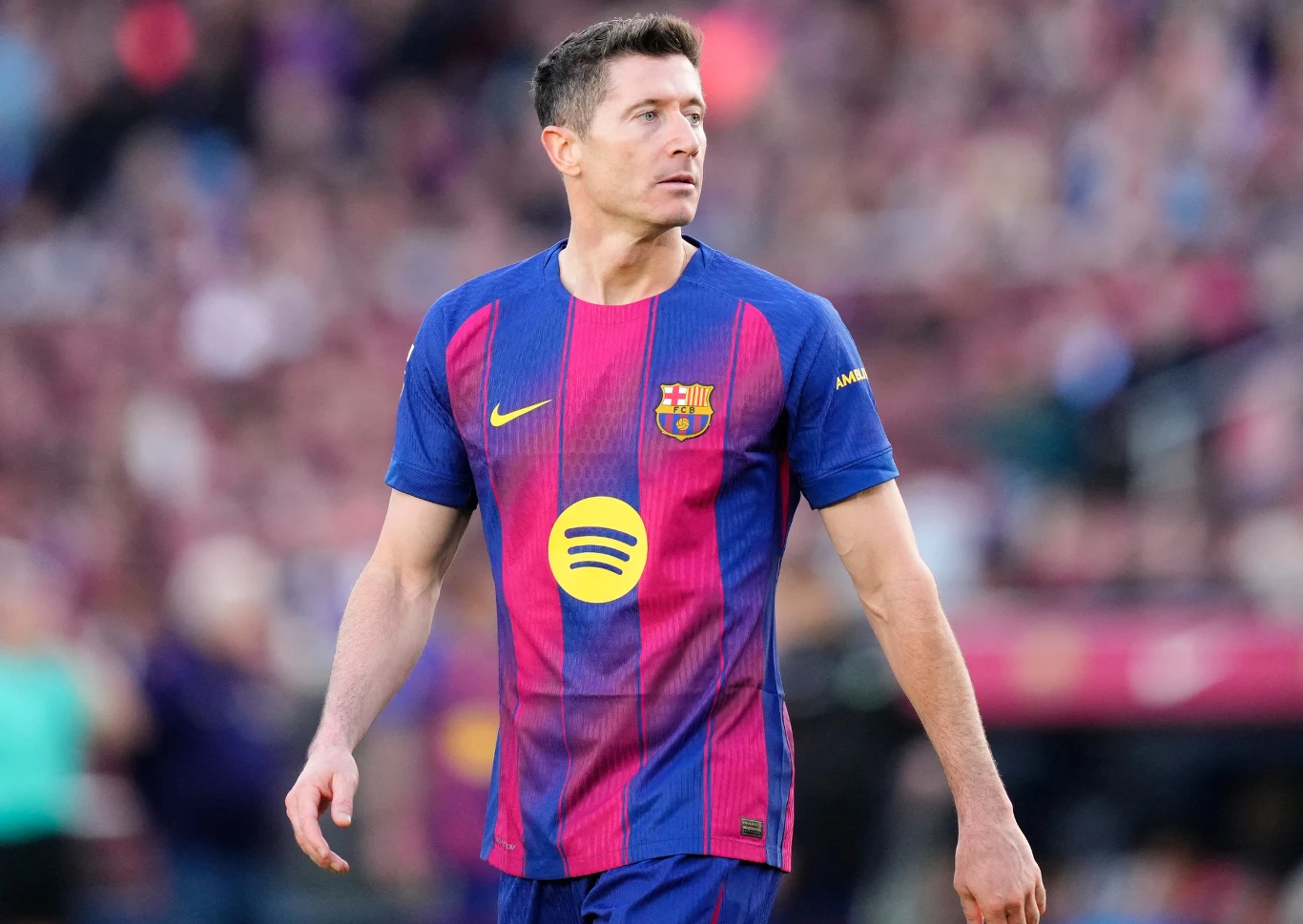 Robert Lewandowski Piłkarz ubrany w granatowo-bordową koszulkę z logo klubu FC Barcelona oraz sponsora, stojący na boisku podczas meczu, w tle rozmyte sylwetki kibiców i stadion.