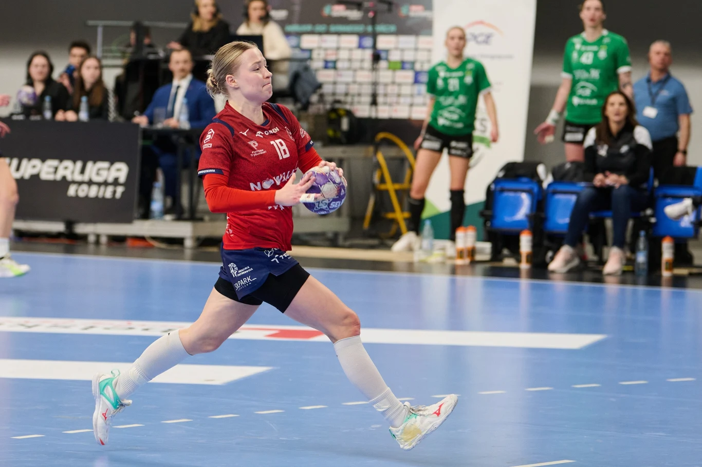 Daria Grobelna Zawodniczka w czerwonej koszulce z piłką w rękach biegnie po parkiecie hali sportowej, w tle widać siedzących zawodników drużyny przeciwnej oraz sędziów i publiczność.