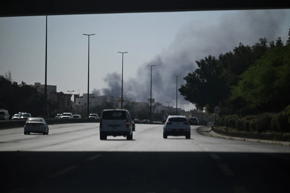Samochody poruszają się po szerokiej, asfaltowej drodze miejskiej, nad którą unoszą się ciemne kłęby dymu pochodzące z odległego obszaru zabudowanego.