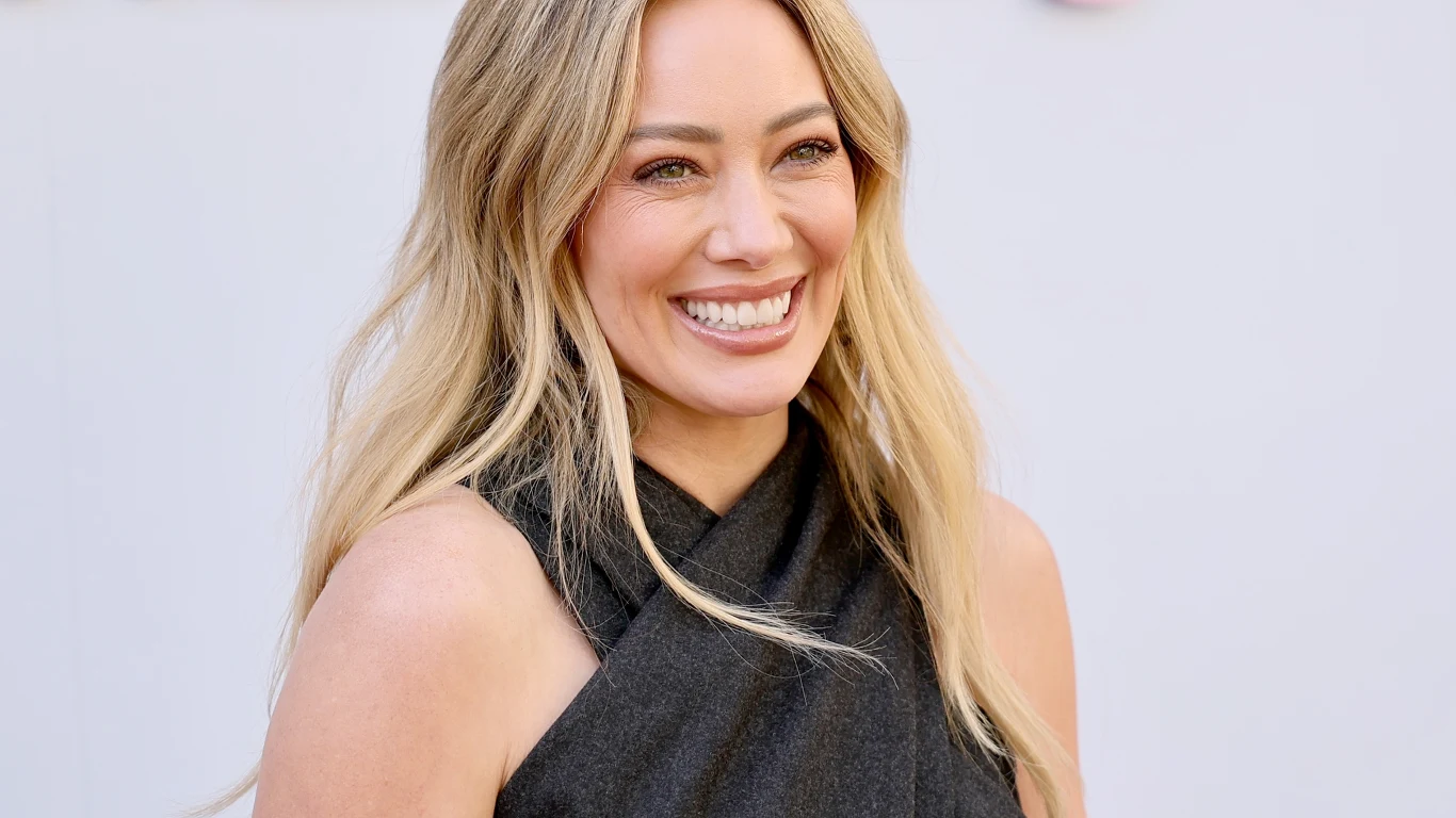 Hilary Duff powraca na muzyczną scenę po 10 latach Uśmiechnięta kobieta o długich blond włosach, ubrana w czarną bezrękawną bluzkę, stoi na jasnym tle.