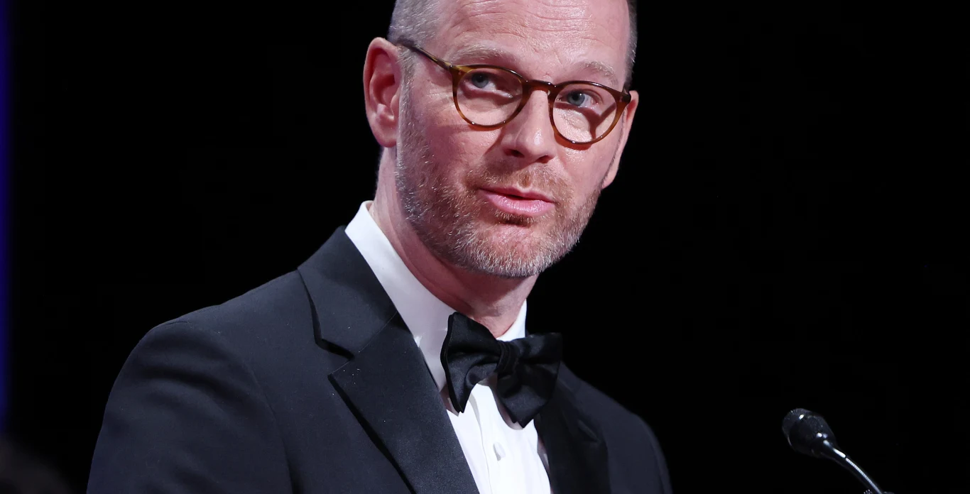 Joachim Trier na festiwalu w Cannes 2025 Mężczyzna w okularach, ubrany w czarny smoking i muszkę, stoi przy mikrofonie i patrzy w stronę widowni, na tle ciemnego tła.