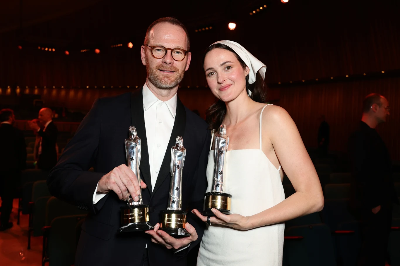 Joachim Trier i Renate Reinsve z Europejskimi Nagrodami Filmowymi za "Wartość sentymentalną" (17 stycznia 2026) Mężczyzna w garniturze oraz kobieta w białej sukni i opasce na głowie trzymają w rękach statuetki nagród, stojąc razem na tle ciemnej sali z rzędami siedzeń.