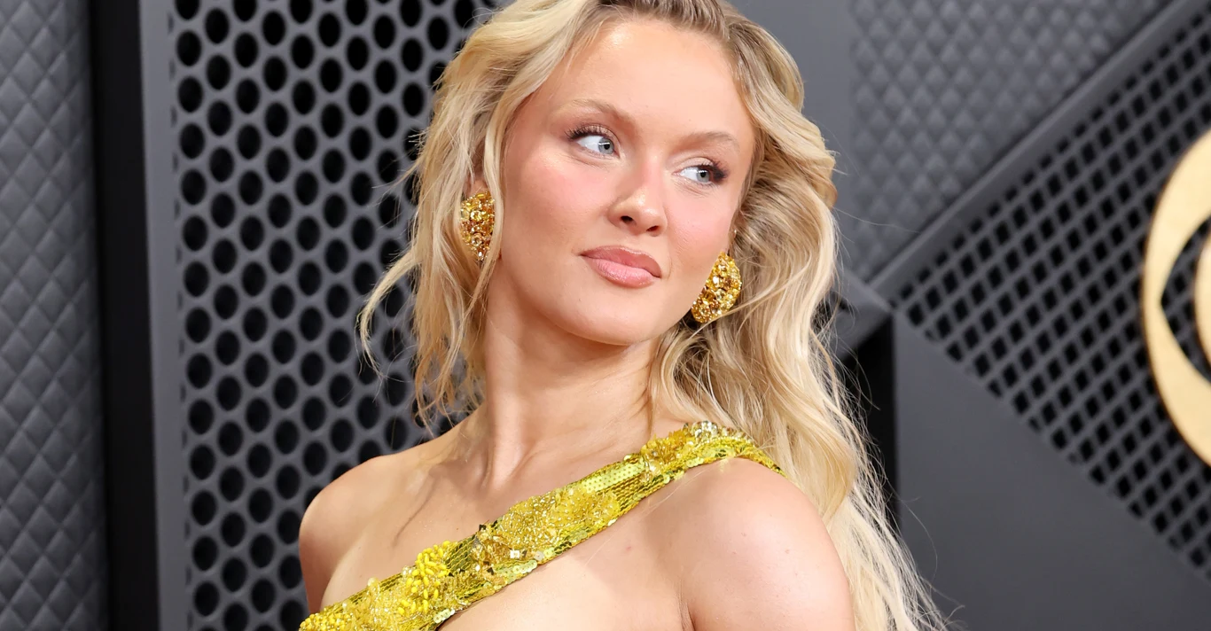 Zara Larsson Młoda kobieta z długimi blond włosami ubrana w elegancką, złotą suknię z efektownymi zdobieniami i złotymi kolczykami, patrząca w bok na tle ciemnej, geometrycznej ściany.