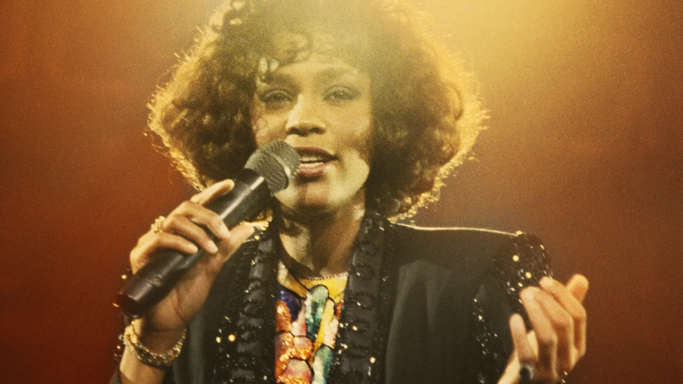 Whitney Houston (1988 r.) Kobieta z mikrofonem na scenie w trakcie śpiewania, ubrana w elegancki czarny żakiet oraz kolorową bluzkę, otoczona ciepłym, scenicznym światłem.