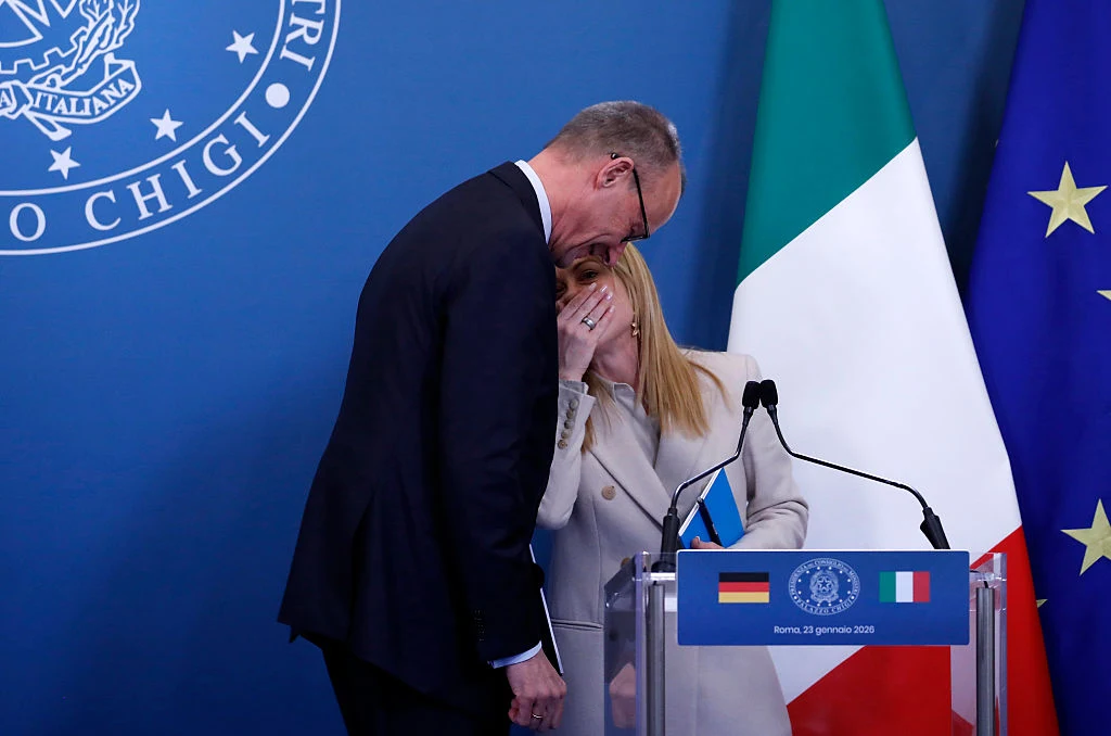 Friedrich Merz i Giorgia Meloni spiskują przeciw ETS Dwoje polityków stoi przy mównicy z mikrofonami, pochylają się do siebie i rozmawiają szeptem. W tle widoczne flagi Unii Europejskiej, Niemiec i Włoch oraz herb włoskiego rządu.