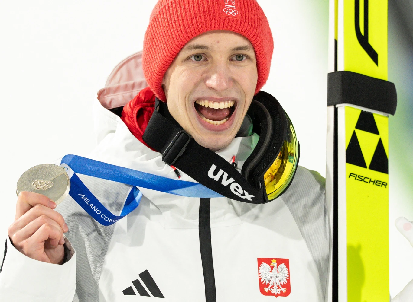 Uśmiechnięty sportowiec w czerwonej czapce i białej kurtce z orzełkiem na piersi, trzymający srebrny medal olimpijski i narty, na szyi widoczna niebieska wstążka z napisem 'Milano Cortina 2026', na ramieniu gogle narciarskie.