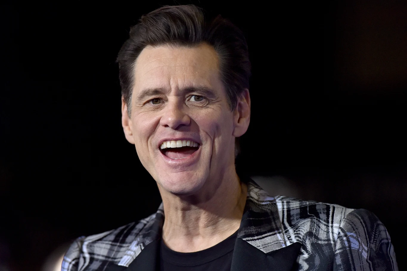 Jim Carrey Mężczyzna w średnim wieku z krótkimi ciemnymi włosami uśmiecha się szeroko, ubrany w marynarkę w biało-czarną kratę na ciemnym tle.