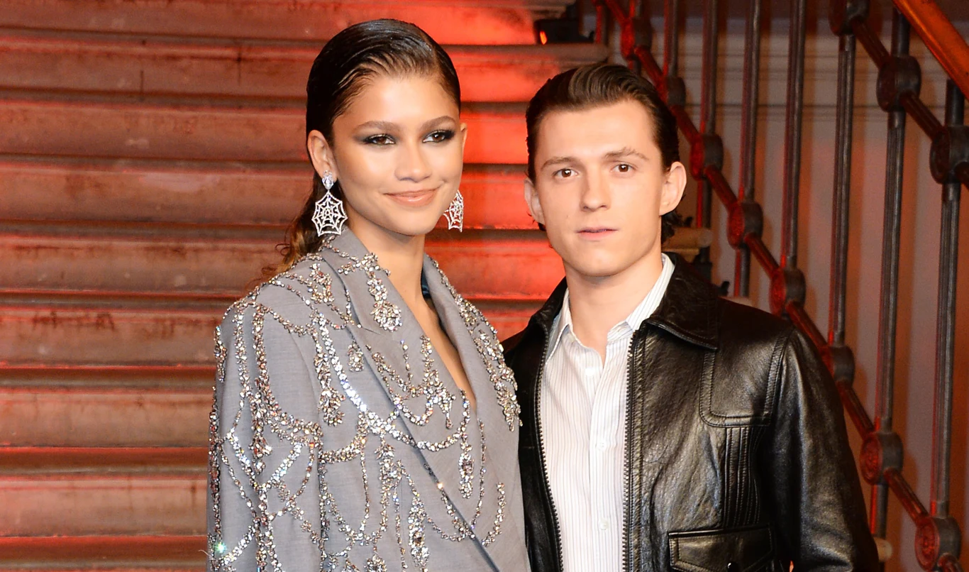 Zendaya i Tom Holland Dwie elegancko ubrane osoby stoją razem na tle ozdobnych schodów. Kobieta w srebrzystym garniturze z błyszczącymi zdobieniami oraz dużymi kolczykami, mężczyzna w czarnej skórzanej kurtce i jasnej koszuli. Oboje patrzą w kierunku aparatu.