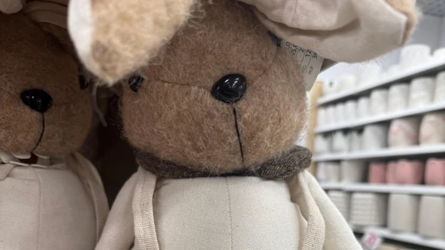 Pluszowy królik ubrany w kremową sukienkę na szelkach z drewnianymi guzikami, stojący na półce w sklepie z artykułami dekoracyjnymi. W tle widoczne podobne zabawki oraz półki wypełnione produktami.