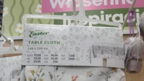 Obrus wielkanocny w kwiatowy wzór z motywami króliczków, zawieszony na wieszaku sklepowym z widoczną etykietą informującą o wymiarach i cenie produktu. W tle fragmenty innych ozdób świątecznych oraz napisy reklamowe.