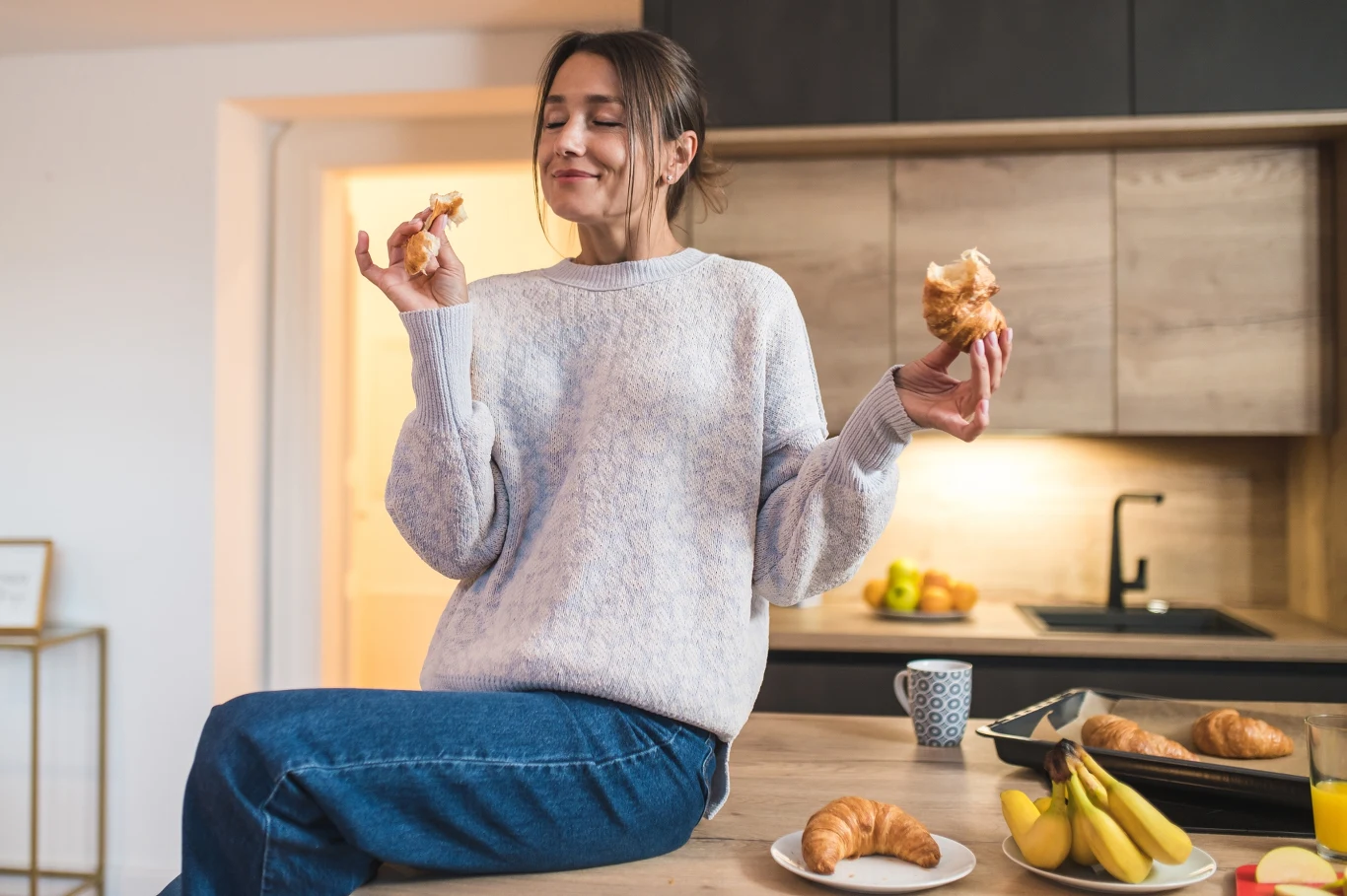Choć gofry czy drożdżówki wydają się być dobrym pomysłem na śniadanie, lepiej postawić na białko Kobieta w swetrze siedzi na blacie kuchennym, trzymając w dłoniach kawałki croissanta i z wyrazem zadowolenia na twarzy. Na blacie znajdują się banany, filiżanka, croissanty oraz naczynia kuchenne.