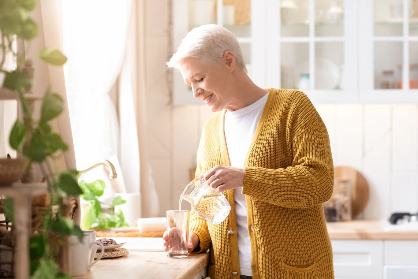 Wypij szklankę wody tuż po przebudzeniu by nawodnić się po nocy Starsza kobieta z krótkimi siwymi włosami ubrana w żółty sweter nalewa wodę z dzbanka do szklanki stojąc w jasnej, przytulnej kuchni otoczonej roślinami.
