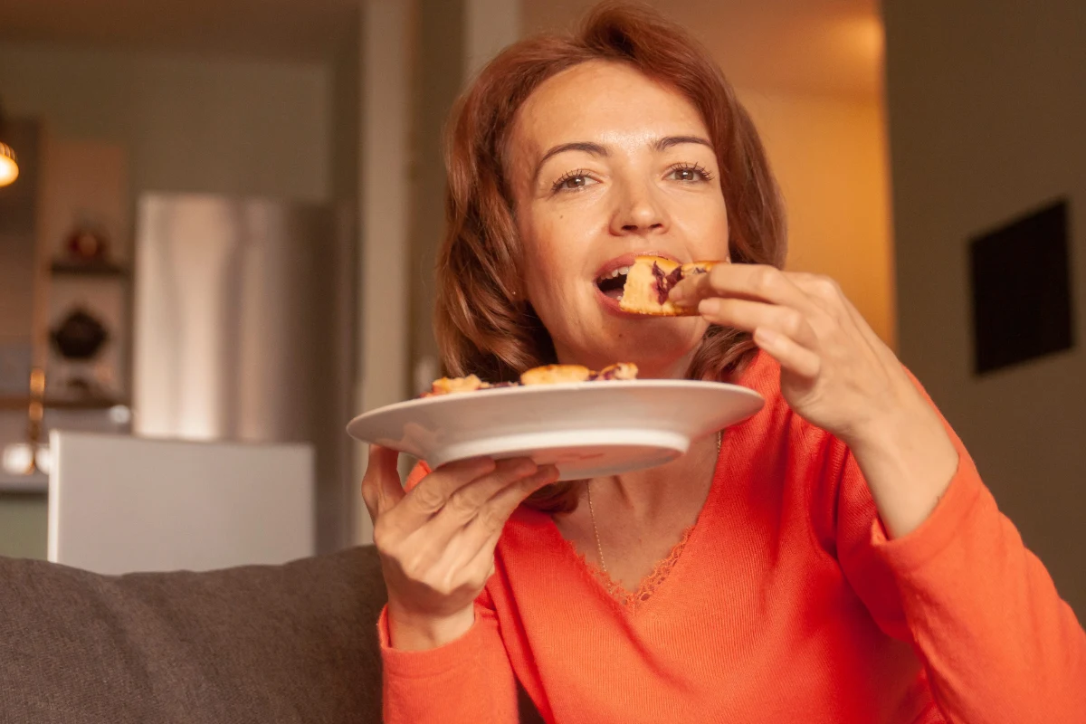 Naukowcy: godzina kolacji ma wpływ na zdrowie i tempo metabolizmu Kobieta w pomarańczowej bluzce siedzi na kanapie i z uśmiechem je pizzę, trzymając talerz w jednej ręce.