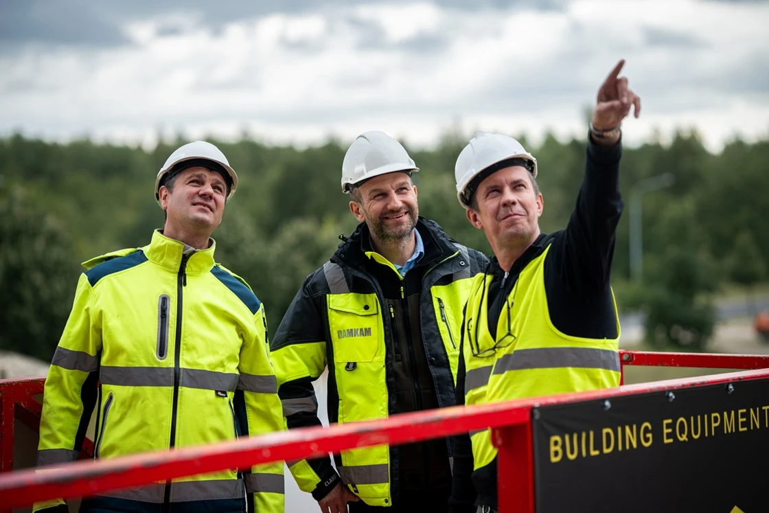 Trzech mężczyzn w odzieży roboczej i kaskach ochronnych stoi na placu budowy, jeden z nich wskazuje ręką coś w oddali. W tle widoczna zielona roślinność oraz sprzęt budowlany.