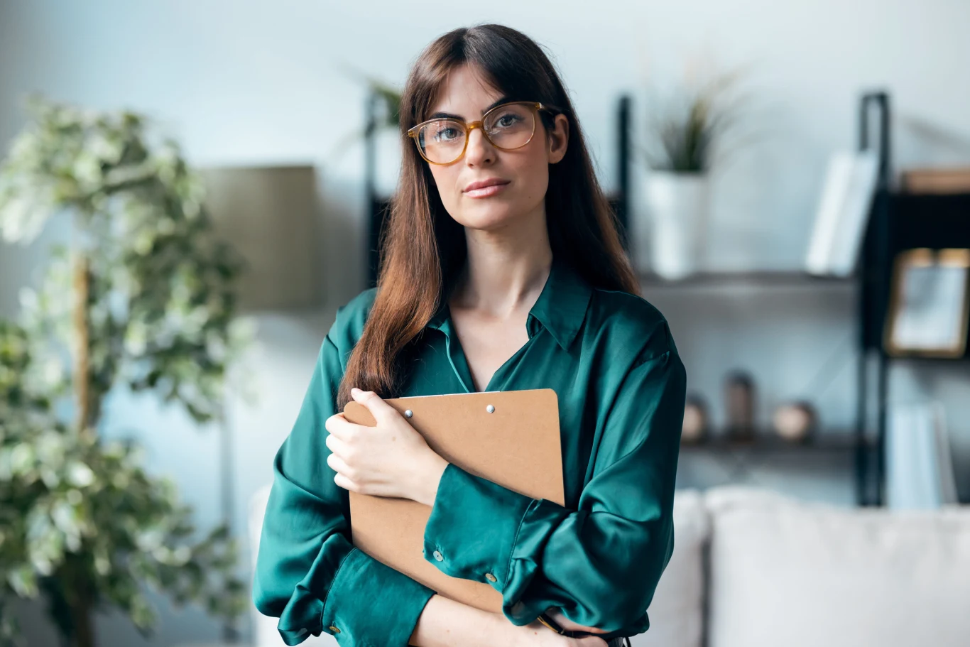Kobieta w okularach o długich brązowych włosach ubrana w zieloną koszulę, trzymająca teczkę w ramionach, stoi w elegancko urządzonym nowoczesnym wnętrzu z rośliną doniczkową i regałem z książkami w tle.