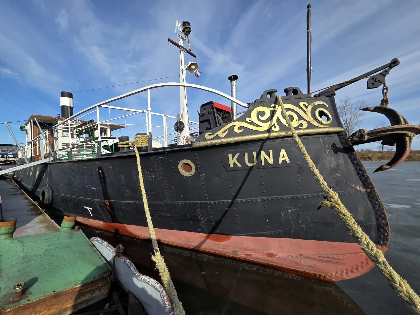 Czarny statek z nazwą KUNA na burcie, ozdobiony złotym ornamentem na dziobie, cumuje przy drewnianym nabrzeżu; na pokładzie widoczny komin i relingi, z przodu masywny żuraw z kotwicą.