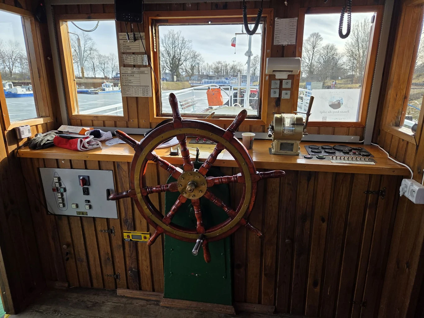 Duże drewniane koło sterowe umieszczone centralnie w kabinie sterowniczej, po lewej stronie panel sterowania z przyciskami, po prawej stronie dźwignia sterownicza i radiotelefon, przez szerokie okna widoczne fragmenty łodzi, woda oraz drzewa w tle.