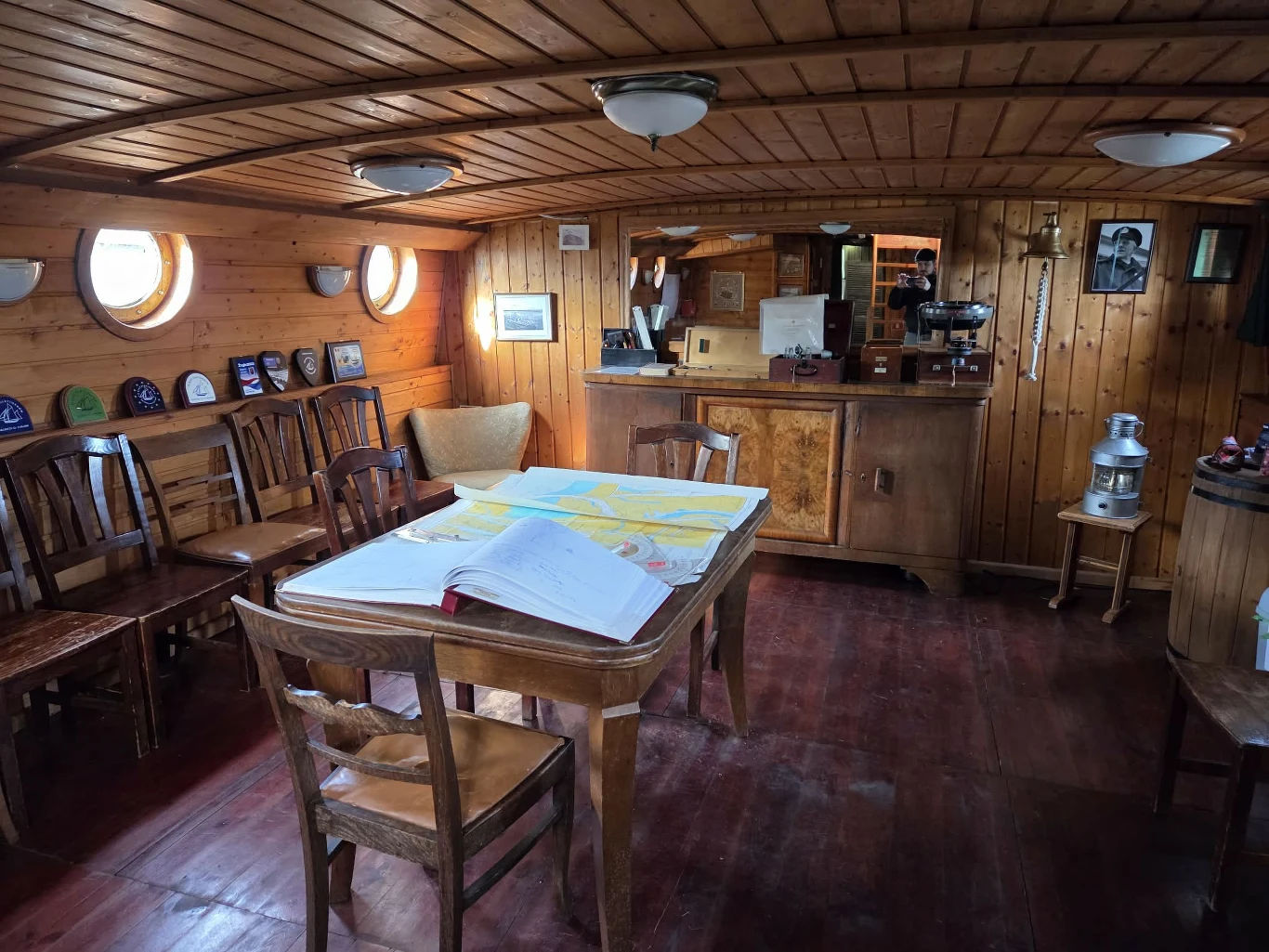 Drewniane wnętrze kajuty statku z centralnie ustawionym stołem, na którym rozłożone są mapy i otwarta księga; wokół stołu ustawione krzesła, na ścianach widoczne bulaje, kolekcja czapek marynarskich, fotografie oraz przyrządy nawigacyjne, a w tle znajd...