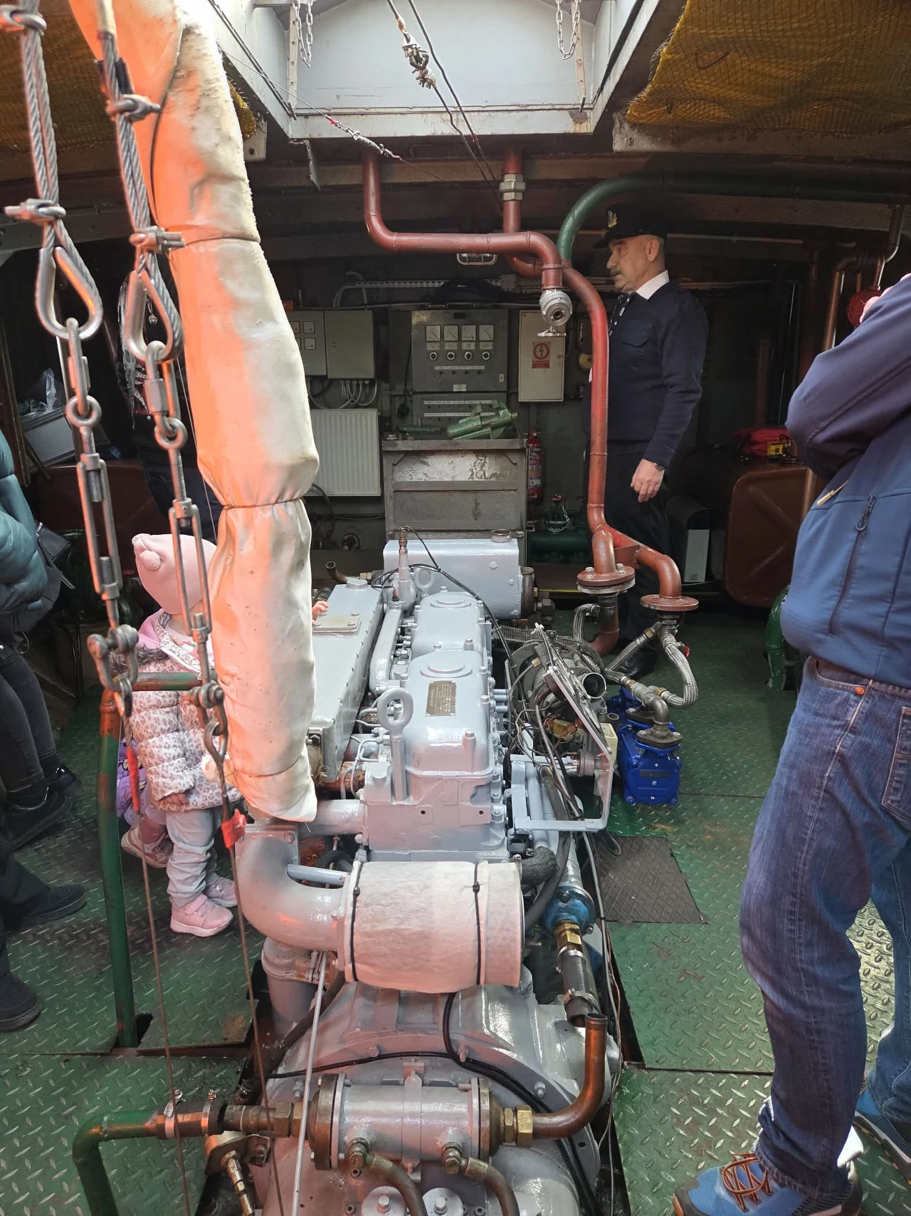 Duży silnik zamontowany na metalowej podłodze, otoczony przez kilka osób, w tle widoczna instalacja elektryczna z tablicą rozdzielczą; mężczyzna w czapce stoi obok instalacji, a po bokach znajdują się inne osoby obserwujące wnętrze maszynowni.