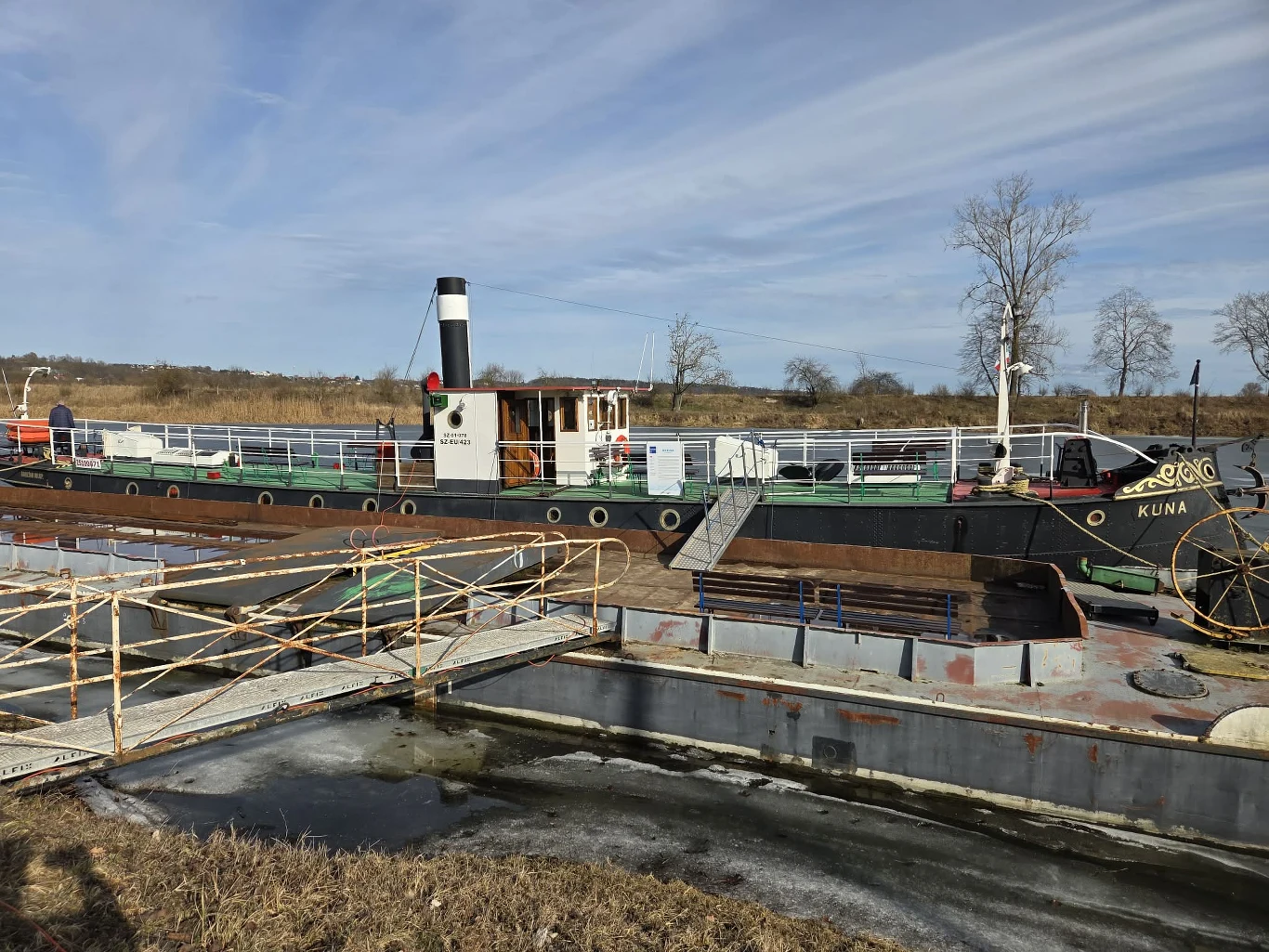 holownik parowy o czarnym i zielonym kadłubie, z białym kominem ozdobionym czarnym pasem oraz nazwą KUNA na dziobie, znajdujący się w śluzie wodnej otoczonej metalowymi balustradami; wokół sucha trawa i pojedyncze drzewa na tle lekko zachmurzonego nieba