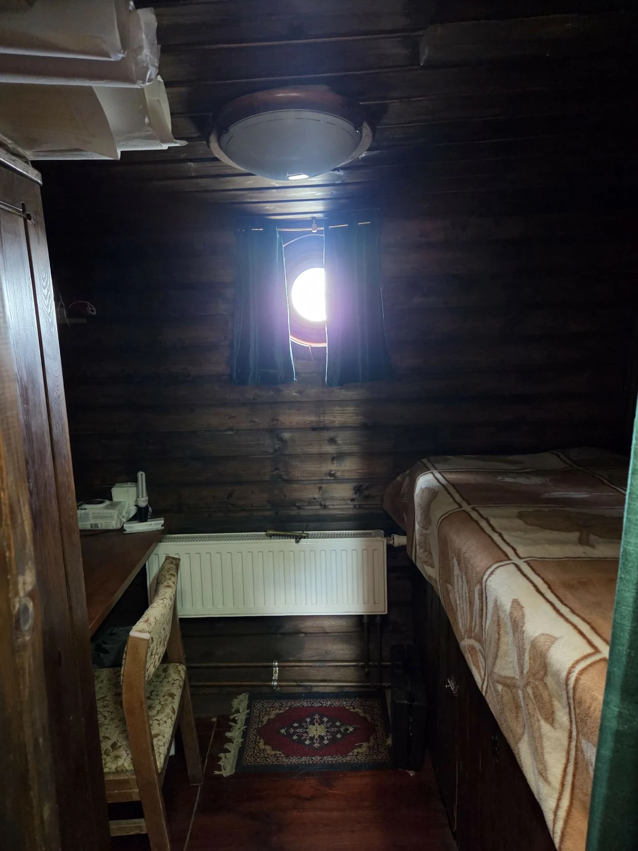 Drewniana izba z wąskim łóżkiem pokrytym wzorzystym kocem, niewielkim stolikiem z krzesłem i okrągłym oknem zasłoniętym ciemnymi zasłonami, przez które wpada światło słoneczne; pod oknem znajduje się biały grzejnik, a na podłodze leży mały dywanik.