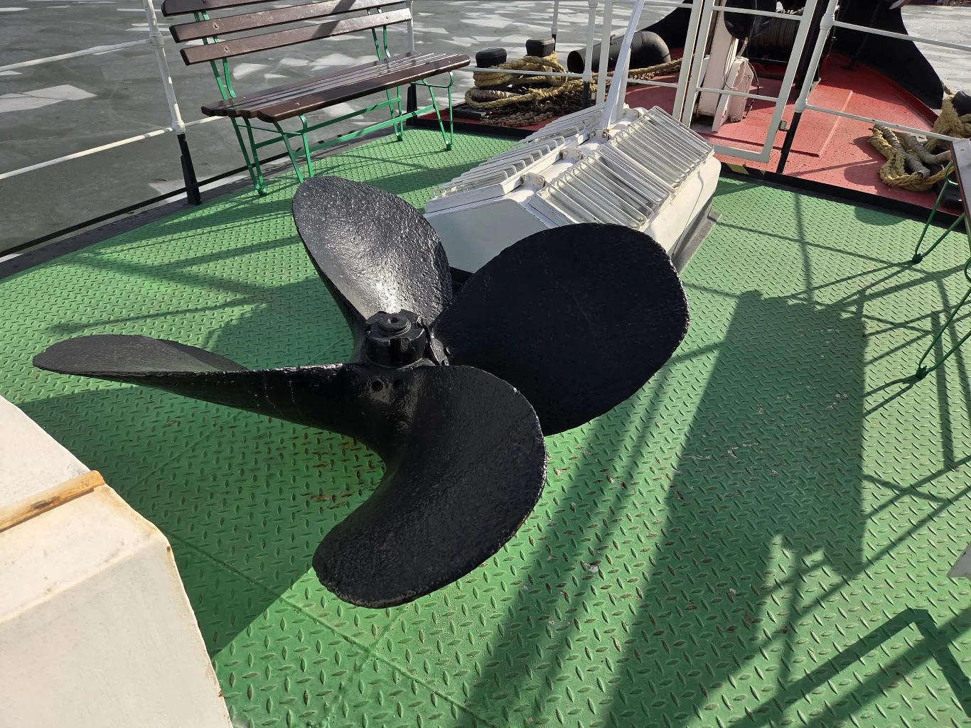 Duża śruba okrętowa o czarnych łopatach ustawiona na zielonej, metalowej podłodze statku; w tle ławka, poręcz i fragmenty pokładu oraz liny marynarskie.