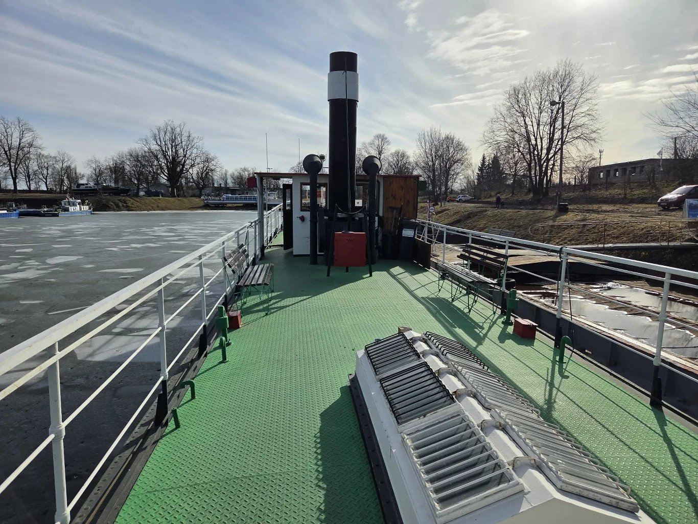 Pokład statku z zieloną podłogą, centralnie ustawiony wysoki komin oraz elementy wyposażenia takie jak ławki i skrzynie, wokół metalowa balustrada, po lewej stronie częściowo zamarznięta rzeka, w tle drzewa bez liści i kilka niskich budynków.