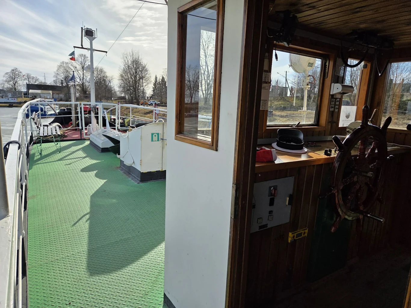 Wnętrze kabiny kapitana statku z widocznym drewnianym kołem sterowym, czapką kapitana na pulpicie oraz widokiem na zielony pokład i relingi prowadzące w stronę dziobu, w tle drzewa bez liści i infrastruktura portowa.