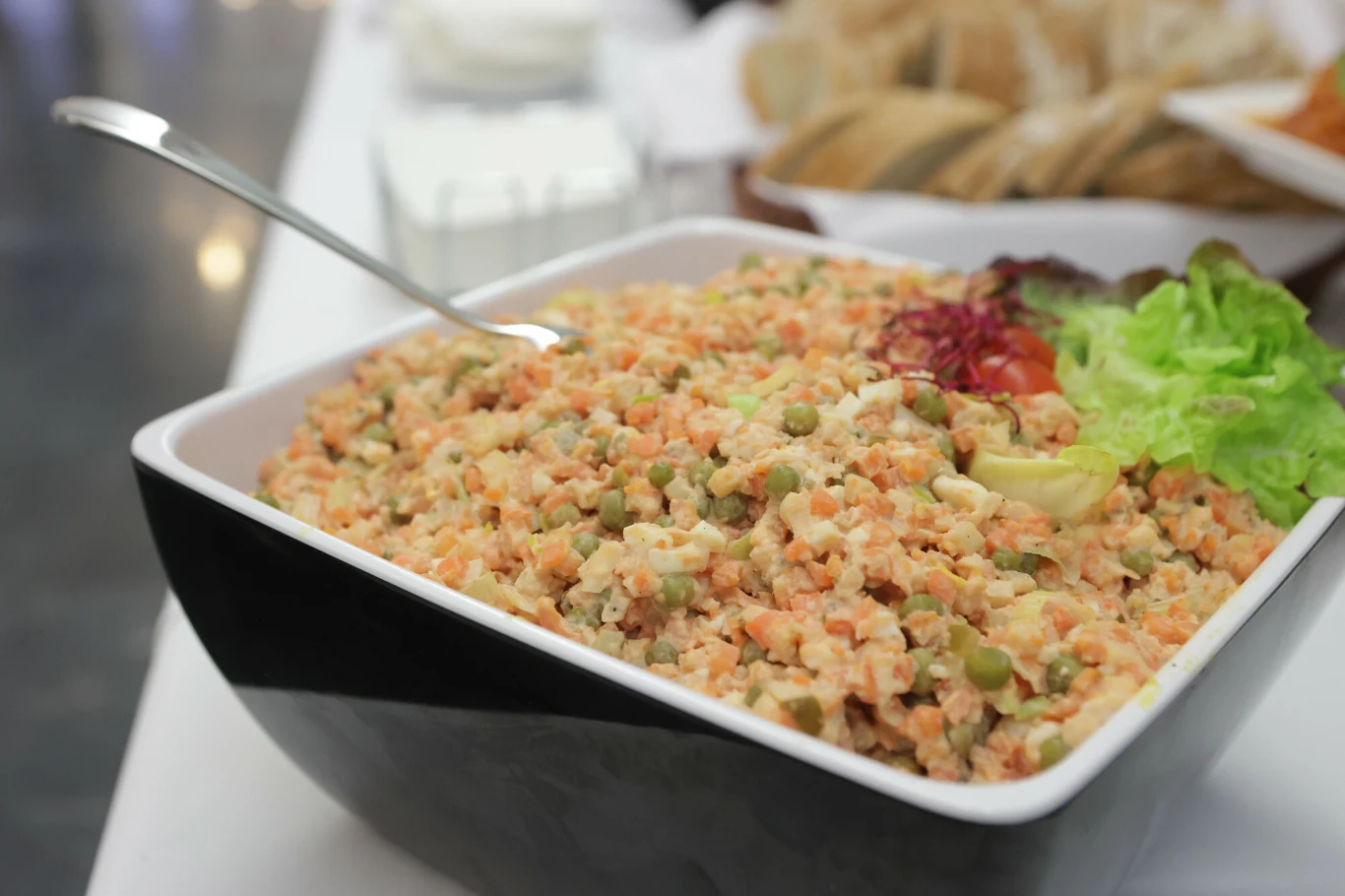 Znudziła ci się już tradycyjna sałatka jarzynowa? Możesz przygotować ją w nieco odmienionym wydaniu Sałatka jarzynowa z groszkiem, marchewką, ziemniakami i majonezem w dużej misce, udekorowana liściem sałaty i plasterkami warzyw, obok leży łyżka do nakładania.