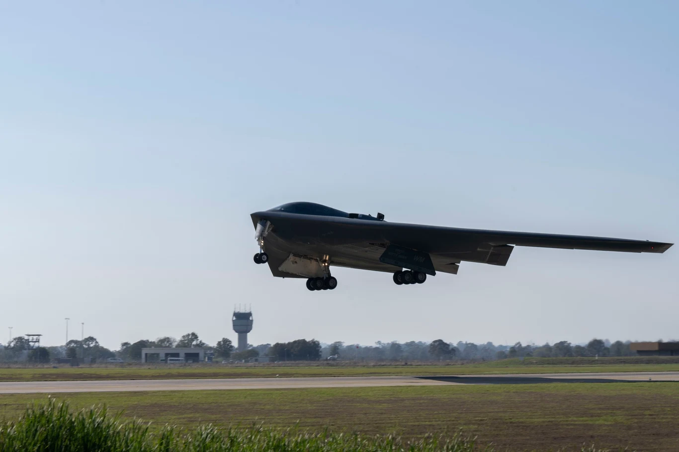 Bombowiec B-2 Spirit Nowoczesny bombowiec stealth unosi się nisko nad pasem startowym, z kołami podwozia opuszczonymi do lądowania; w tle widoczne są budynki lotniska i wieża kontroli lotów.