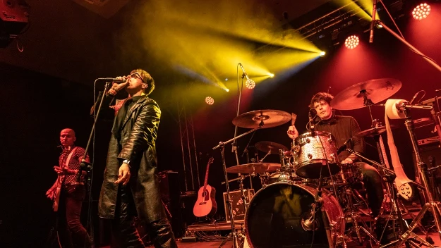 Zespół muzyczny w trakcie koncertu, wokalista w skórzanym płaszczu śpiewa do mikrofonu, obok niego gitarzysta, perkusista gra energicznie na bębnach, scena oświetlona żółtymi i pomarańczowymi reflektorami, w tle gitara akustyczna oparta o stojak.