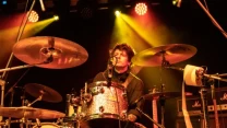 Perkusista gra na scenie, otoczony zestawem bębnów i talerzy, z mikrofonem przed twarzą oraz żółtym światłem reflektorów skierowanym w jego stronę, po prawej stronie widoczne wzmacniacze gitarowe.