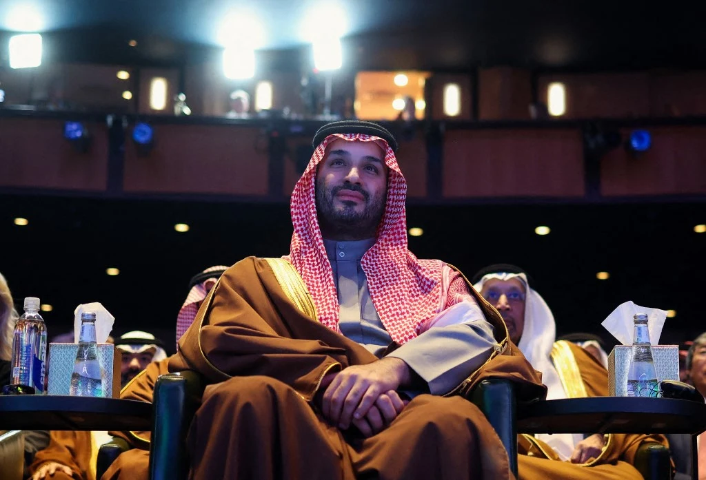 Mężczyzna w tradycyjnym arabskim stroju siedzi na fotelu na tle sali widowiskowej, wokół niego widoczne są butelki wody oraz inni uczestnicy wydarzenia.