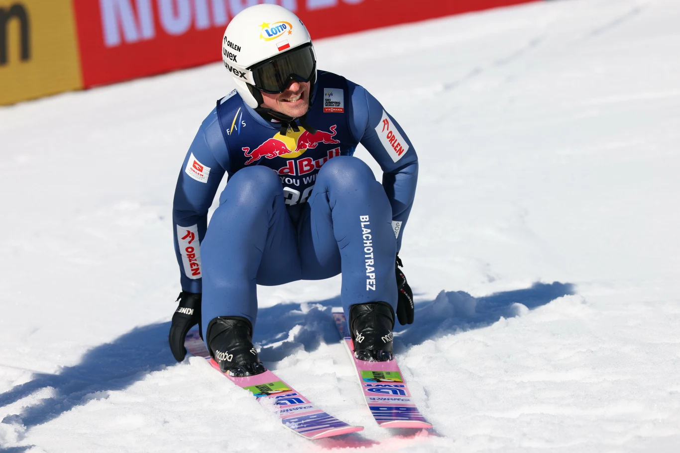 Skoczek narciarski w niebieskim kombinezonie i kasku z widocznymi logotypami sponsorów, siedzi na śniegu po wylądowaniu skoku, trzymając narty równolegle przed sobą.