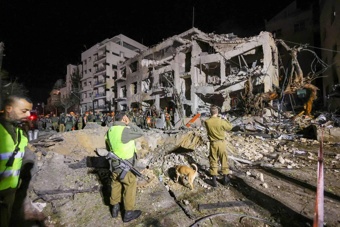 Iran nie pozostaje bierny. Na zdjęciu skutki bombardowań w Tel Awiwu Zrujnowany budynek po eksplozji w mieście, wokół którego pracują służby ratownicze w odblaskowych kamizelkach oraz psy ratownicze przeszukujące gruzowisko.