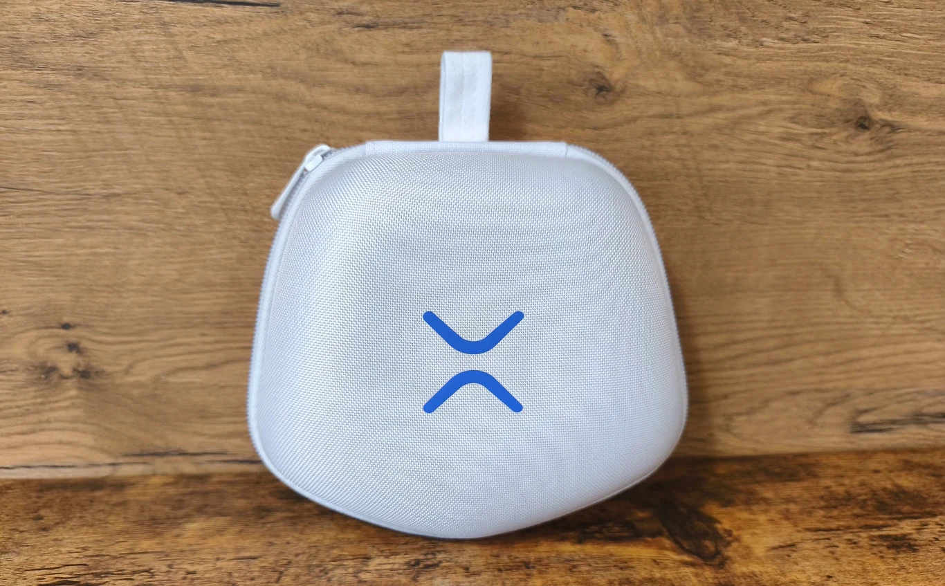 Biała, półokrągła saszetka z niebieskim logo umieszczonym centralnie, zamykana na zamek błyskawiczny, ustawiona na drewnianej powierzchni na tle drewnianej ściany.