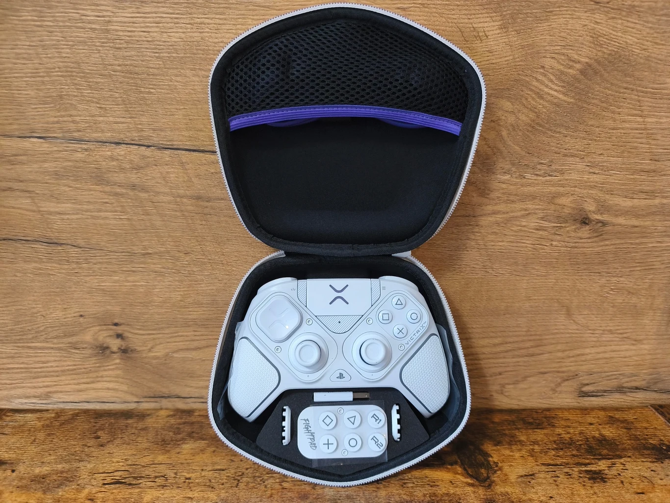 Biały kontroler do gier zamknięty w dopasowanym etui ochronnym, ułożony na drewnianej powierzchni, centralnie skupione elementy sterujące i przyciski wyróżniające się na tle czarnego wnętrza opakowania.