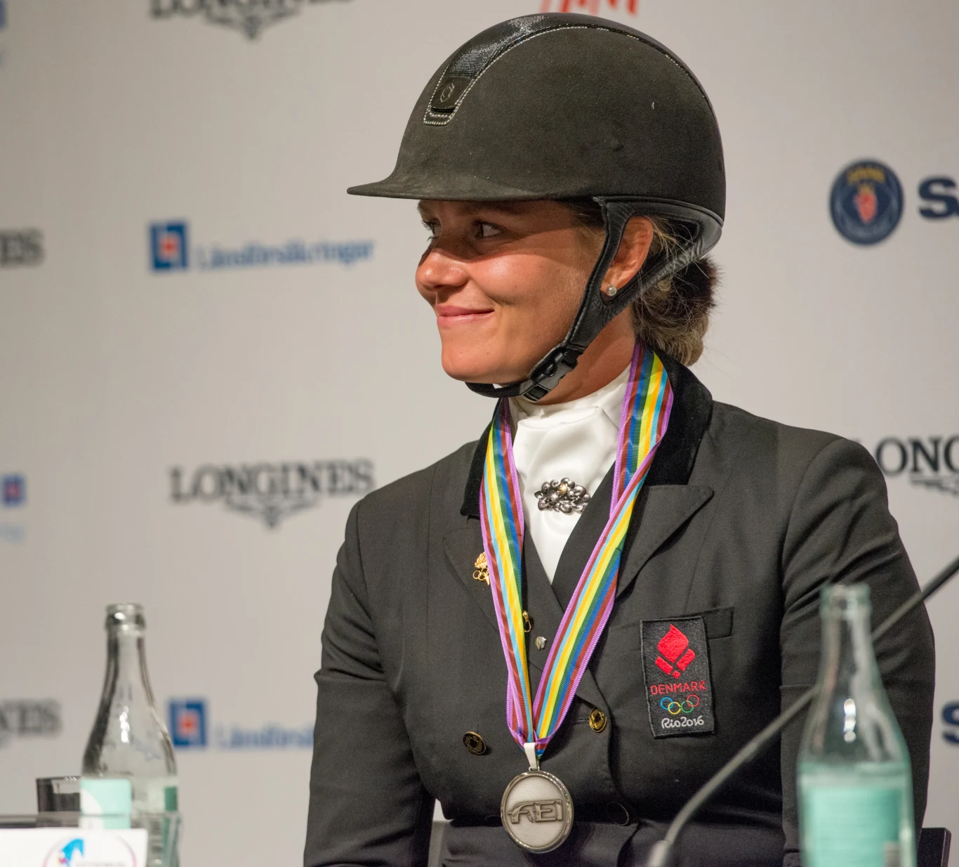 Kobieta w stroju jeździeckim i kasku siedzi przy stole, na szyi ma srebrny medal zawieszony na kolorowej wstążce, za nią widoczne logotypy sponsorów na ścianie.