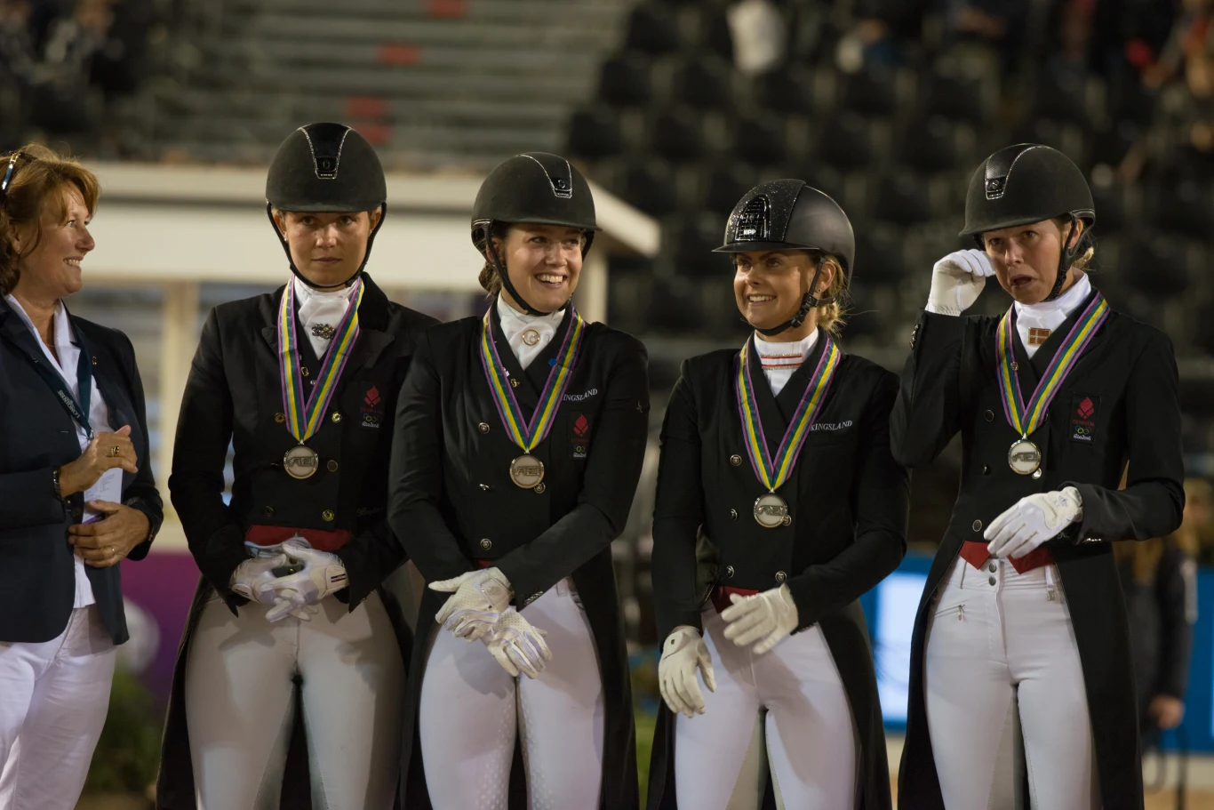 Cztery kobiety w strojach jeździeckich z kaskami i medalami na szyjach stoją razem, uśmiechając się i rozmawiając, obok nich stoi kobieta w formalnym stroju.