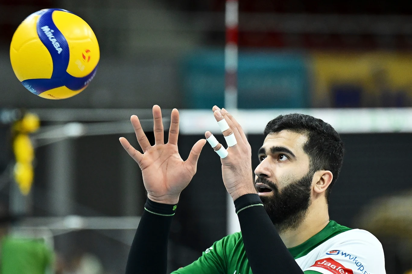 Amirhossein Esfandiar, przyjmujący InPost ChKS Chełm Siatkarz w zielono-białym stroju koncentruje się na piłce lecącej w jego kierunku, unosząc ręce do przyjęcia, z opatrzonymi palcami lewej dłoni, w tle widoczne rozmyte elementy hali sportowej.